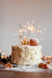 a cake with white frosting and white chocolate, with a sparkler and a mandarin on top